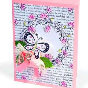 May include: A handmade greeting card with a pink background featuring a butterfly and floral design. The butterfly has black and white wings with yellow accents. A pink ribbon and lace add a decorative touch. The card has text from a dictionary page.
