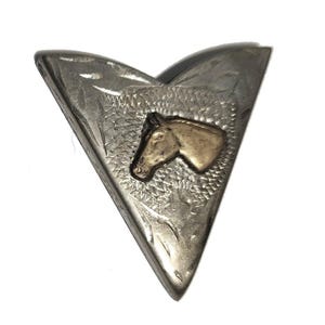 May include: A silver-toned, V-shaped metal object with a gold-colored horse head in the center. The metal has an etched design around the horse head and along the edges. The background is white.