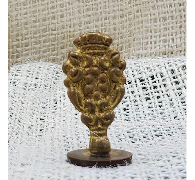 以下が含まれることがあります： 王冠と花のモチーフが装飾された金色の金属製の印鑑。印鑑は丸い台座に載せられています。