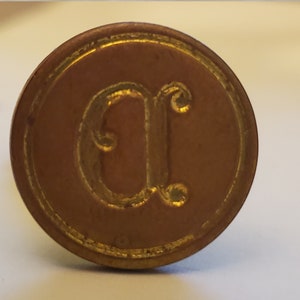 以下が含まれることがあります： 中央に浮き彫りの文字「C」が刻まれた金色の金属製の印鑑。印鑑は円形のデザインで、浮き彫りの縁取りが施されています。