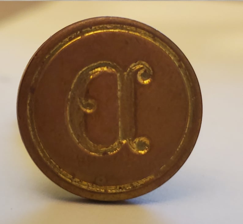 以下が含まれることがあります： 中央に浮き彫りの文字「C」が刻まれた金色の金属製の印鑑。印鑑は円形のデザインで、浮き彫りの縁取りが施されています。