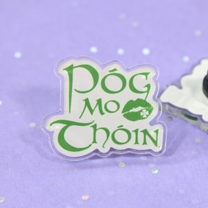 May include: A clear acrylic pin with a green shamrock and the text "Dóg Mo Chóin" in green lettering. The pin has a black metal backing.