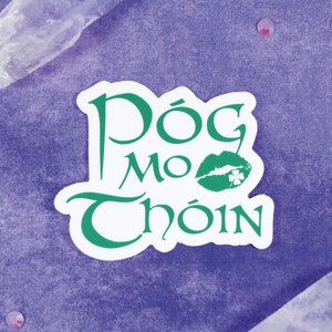 May include: A white sticker with green text and a green shamrock-shaped lip print. The text reads "Dóg Mo Chóin".