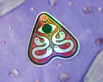 May include: A holographic sticker in a triangular shape, featuring a design with a large eye, a crescent moon, stars, and several ghost figures. The sticker has a red and rainbow gradient with a black border. The sticker is on a purple surface.