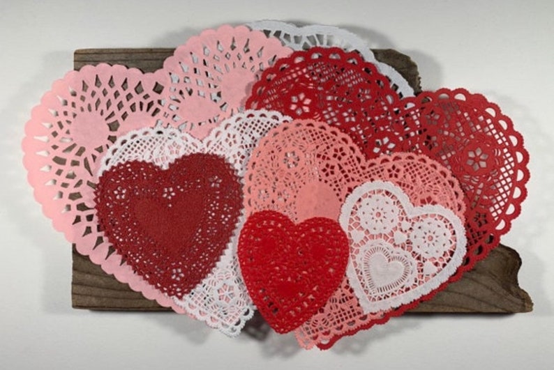 Peut inclure: Une collection de doilies en papier rouge, rose et blanc en forme de c&oelig;ur. Les doilies sont dispos&eacute;s en forme de c&oelig;ur sur une surface en bois.
