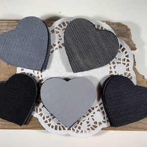 May include: Five felt heart shapes in various shades of gray, black, and white. The hearts are arranged on a white doily and a piece of wood.