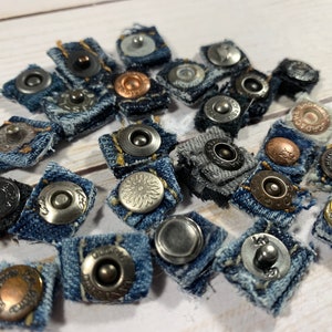 May include: A collection of denim-covered snap buttons with various metal finishes, including silver, bronze, and black. The buttons are attached to small, frayed denim squares, creating a rustic, handcrafted appearance. Ideal for sewing and crafting projects.