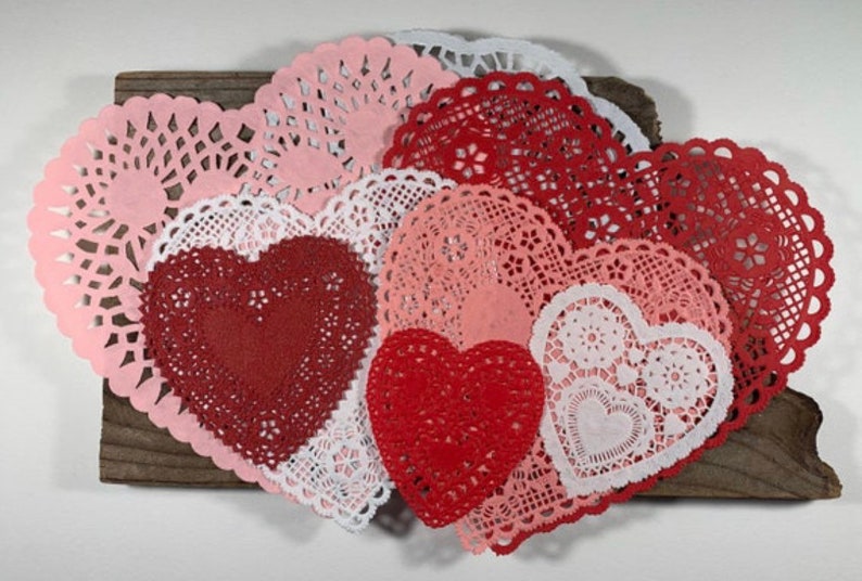 Peut inclure: Une collection de doilies en papier rouge, rose et blanc en forme de c&oelig;ur. Les doilies sont dispos&eacute;s en forme de c&oelig;ur sur un morceau de bois vieilli.