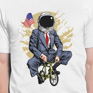 Puede incluir: Camiseta blanca con un gráfico de un astronauta con un traje montando una bicicleta. El astronauta sostiene una bandera estadounidense. El gráfico está sobre un fondo blanco con nubes.