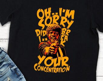 May include: Black t-shirt featuring a stylized image of a man pointing a gun, with the text "OH...I'M SORRY DID I BREAK YOUR CONCENTRATION" in orange. The graphic is a pop culture reference. The shirt is paired with blue jeans.