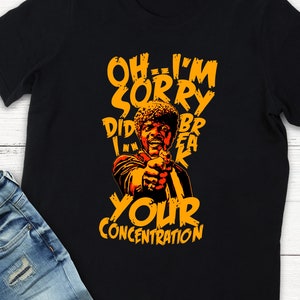 May include: Black t-shirt with an orange graphic of a man holding a gun, and the text "OH...I'M SORRY DID I BREAK YOUR CONCENTRATION". The design is a pop culture reference. The shirt is displayed with blue jeans.