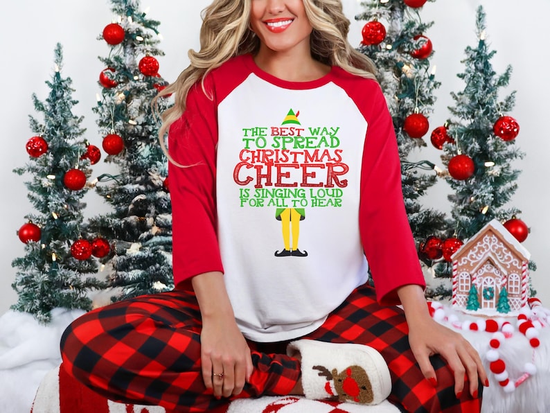以下が含まれることがあります： 赤い袖とクリスマスのエルフのグラフィックが付いた白い長袖シャツ。シャツには「The best way to spread Christmas cheer is singing loud for all to hear」というテキストが書かれています。