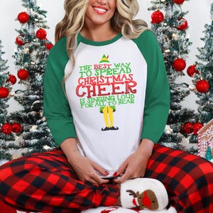 以下が含まれることがあります： 白と緑のロングスリーブシャツに、クリスマスエルフのグラフィックと「The best way to spread Christmas cheer is singing loud for all to hear」というテキストがプリントされています。この人は赤と黒のチェック柄のパジャマパンツを履いています。