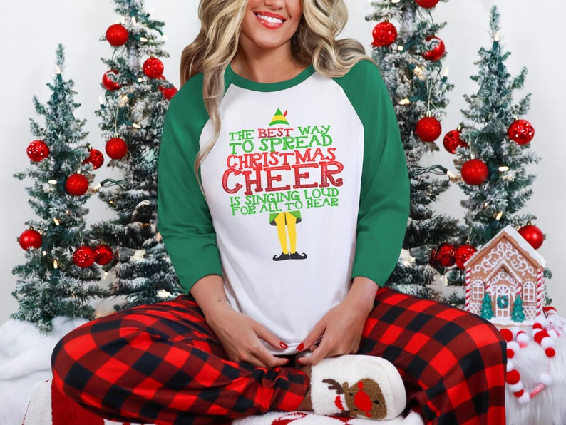 以下が含まれることがあります： 白と緑のロングスリーブシャツに、クリスマスエルフのグラフィックと「The best way to spread Christmas cheer is singing loud for all to hear」というテキストがプリントされています。この人は赤と黒のチェック柄のパジャマパンツを履いています。