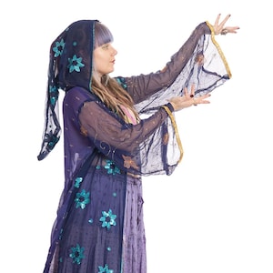 Peut inclure: Un ensemble fluide et superposé dans les tons de violet et bleu marine. La tenue comprend une cape à capuche avec des ornements floraux et une longue jupe. Les manches ont une bordure dorée. La personne porte des bottes noires.
