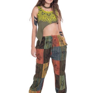 May include: An olive green sleeveless crop top featuring a yellow mandala print on the chest, paired with loose-fitting patchwork trousers in various earth tones including red, green, and brown, decorated with black printed symbols and patterns.