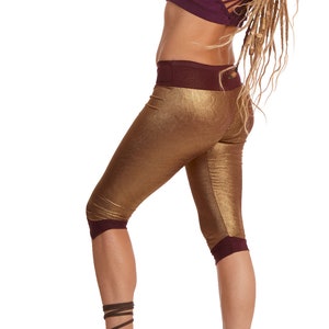 Puede incluir: Una mujer con un top morado, leggings dorados con una cintura morada y sandalias marrones. Lleva un sombrero de copa rosa con una flor y tiene rastas largas.