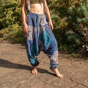 Op de afbeelding: Een persoon draagt een groene crop top, een vest met patroon en een blauwe harembroek met een trekkoord in de taille. De broek heeft lappen blauwe, turquoise en grijze stof. De persoon staat op een stenen pad in een tuin.