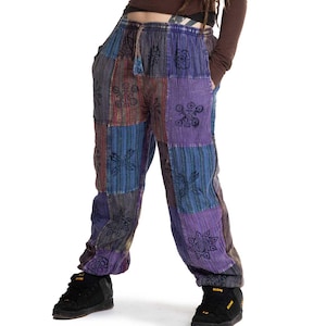 Puede incluir: Una persona con una camiseta marrón de manga larga y pantalones patchwork. Los pantalones son una mezcla de cuadrados azules, morados y marrones con diseños florales. Zapatillas negras completan el look.