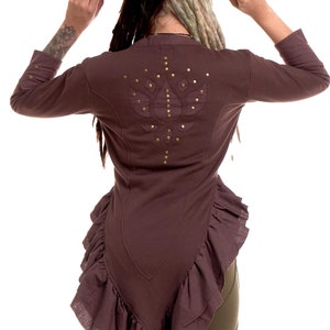 May include: A brown, long-sleeved top with a ruffled hem and a lotus flower design on the back. The top is adorned with gold-colored studs. The model is wearing a black fedora hat.