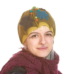 Puede incluir: Una mujer con un sombrero de fieltro verde adornado con hojas y cuentas, una bufanda de ganchillo burdeos y una camisa azul oscuro.