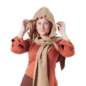 May include: A woman wearing a brown hooded tunic with a long brown scarf. The tunic has a long, pointed hem and the scarf is tied around her neck. She is wearing a brown skirt and has long, braided hair.