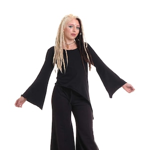 Puede incluir: Una mujer con un top negro de manga larga con mangas acampanadas y pantalones negros de pierna ancha. Tiene rastas rubias y marrones y lleva un collar plateado.