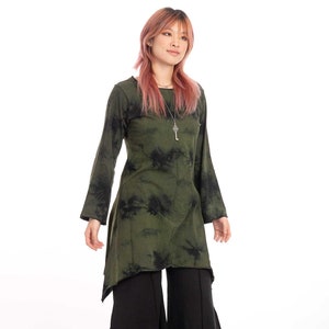 Puede incluir: Una mujer con una túnica de manga larga de color verde y negro con estampado tie-dye, un collar con un colgante de llave y pantalones negros de pierna ancha.