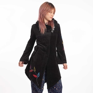 Peut inclure: Une veste en velours noir avec une longue ligne d'ourlet asymétrique et des feuilles brodées colorées. La veste a une capuche et une fermeture à bouton. Le mannequin porte un pantalon bleu à motifs paisley et des bottes noires.