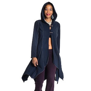 Pode incluir: Uma mulher vestindo uma jaqueta azul escura com capuz e uma bainha longa e assimétrica, e uma calça roxa escura com uma bainha franjada. Ela está usando botas pretas.