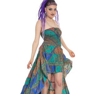 May include: A woman with purple hair is wearing a blue, green, and orange patterned dress with a high-low hemline. The dress has a ruffled skirt and a fitted bodice. She is wearing silver platform sandals.