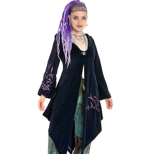 Può includere: Una donna indossa una lunga giacca di velluto blu scuro con ricami floreali viola. La giacca ha un cappuccio e una chiusura con un solo bottone. Indossa anche pantaloni a fantasia verde e nero e stivali neri.