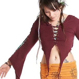 May include: A woman wearing a maroon long-sleeved top with a lace-up front and a yellow and orange skirt with a sun design. The top is cropped and has a frayed hem. The skirt is long and flowy.