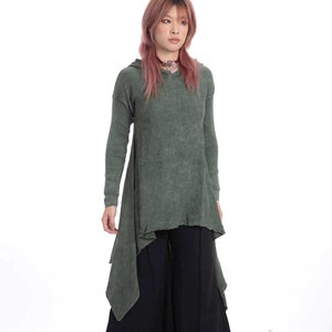 以下が含まれることがあります： 緑色のフード付きセーターと黒いワイドレッグパンツを着用した女性。セーターは裾が長く、左右非対称になっている。