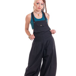 Op de afbeelding: Een vrouw met dreadlocks en kleurrijke make-up draagt een zwarte jumpsuit met daaronder een turquoise tanktop. Ze draagt zwarte en oranje sneakers.