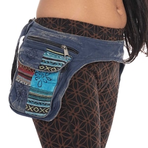 Puede incluir: Una riñonera de mezclilla azul con cierre de cremallera y varios bolsillos. Los bolsillos están decorados con patrones bordados coloridos. La riñonera se lleva alrededor de la cintura de una persona que lleva pantalones de yoga marrones con estampado.