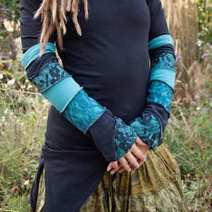 Puede incluir: Una camisa negra de manga larga con hombros descubiertos con ribetes de encaje azul verdoso y negro en las mangas. La camisa se lleva con una falda estampada verde y dorada.