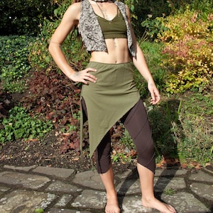 May include: A woman wearing a green and brown outfit with a patterned vest. She is standing outdoors on a stone path.
