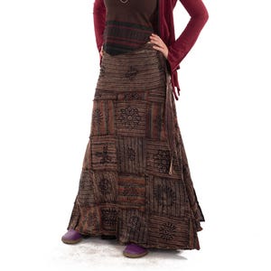 May include: A woman in a layered outfit. A burgundy hooded jacket over a brown tank top with black and red stripes. A long, patchwork skirt with floral designs and purple shoes complete the look. The outfit has a bohemian style.