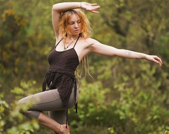 May include: A woman wearing a brown crocheted top and brown leggings poses in a forest setting. She has long, blonde hair and is wearing a necklace with a pendant.