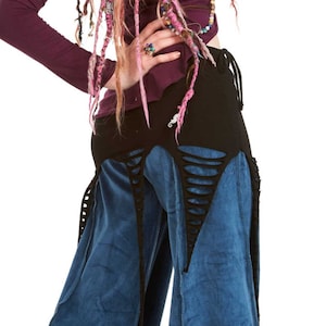 Op de afbeelding: Een persoon draagt een donkerpaarse top met lange mouwen en een blauwe fluwelen broek met een zwarte rok erover. Haar roze haar is gestyled met dreadlocks en een blauwe bloem. De outfit heeft een bohemien stijl.