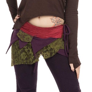 May include: A woman with short, pink hair is wearing a brown, long-sleeved shirt with a high-low hemline. She is also wearing a multi-layered skirt with a green, purple, and red color scheme. The skirt is tied at the waist with a red sash.