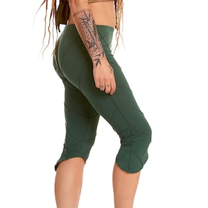 May include: A woman wearing a brown vest with decorative beading, green capri pants, and brown leather sandals. The vest has a halter neck and button closure. The capri pants have a unique seam design. The sandals have multiple straps.