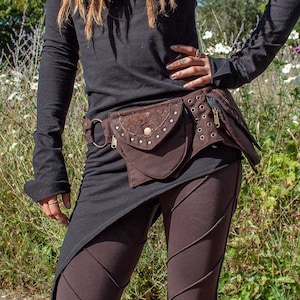 Puede incluir: Una persona vestida con una camiseta negra de manga larga, leggings marrones y una riñonera marrón. La riñonera tiene una solapa decorativa y está adornada con tachuelas metálicas. El atuendo está diseñado en un estilo alternativo.