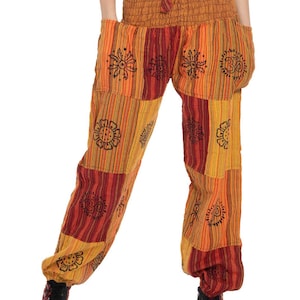 Könnte beinhalten: Weit geschnittene Hose mit gesmoktem Gummibund und Kordelzug. Der Stoff zeigt ein Patchwork-Muster aus vertikalen Streifen in Orange-, Gelb- und Rottönen, akzentuiert durch schwarze kreisförmige Blumen- und Om-Symbol-Aufdrucke.