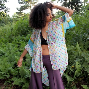 May include: A woman wearing a white, blue, and yellow patterned kimono with a purple pair of wide-leg pants. She has long, curly hair and is standing in a forest setting.