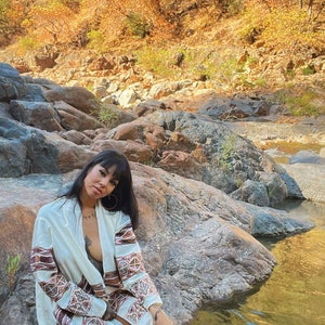 以下が含まれることがあります： 白と茶色の模様のカーディガンを着た女性が、小川沿いの岩の上に座っています。小川は岩と紅葉した木々に囲まれています。
