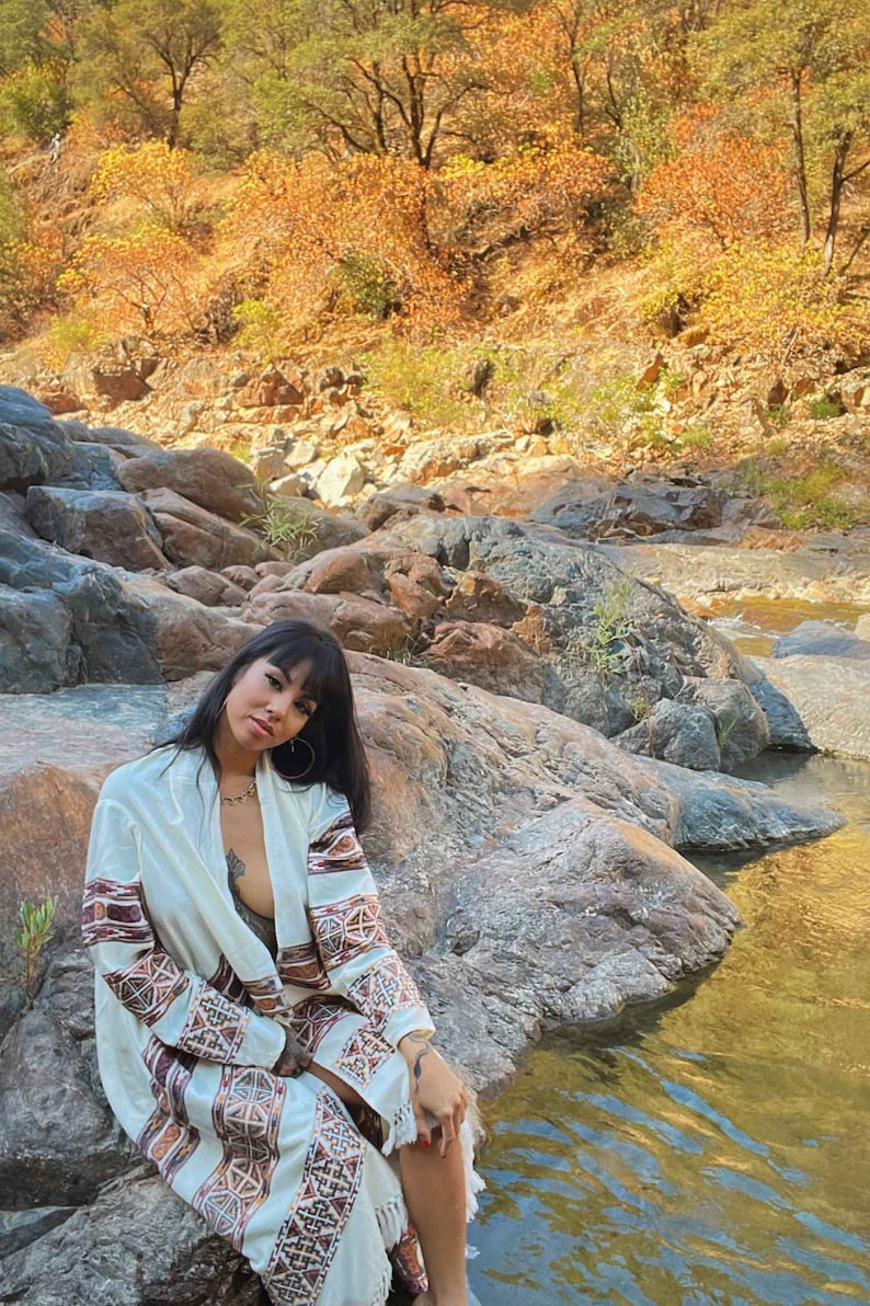 以下が含まれることがあります： 白と茶色の模様のカーディガンを着た女性が、小川沿いの岩の上に座っています。小川は岩と紅葉した木々に囲まれています。