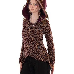 May include: A long-sleeved, hooded jacket in a deep burgundy hue, featuring an intricate gold and brown paisley pattern. The garment includes a large hood with decorative fringe trim, a wrap-style front closure secured by two dark buttons, and a high-low hemline.
