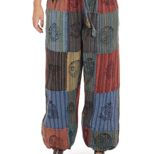 Op de afbeelding: Losvallende patchwork broek met een veelkleurig gestreept patroon in rood-, blauw- en groentinten. De stof is versierd met zwarte gedrukte symbolen, waaronder Om- en Ganesha-motieven. De broek heeft een elastische tailleband met een trekkoord.